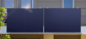 Two large solar panels are mounted on the balcony of a modern building. The panels are positioned side by side and face outward, presumably for optimal sunlight absorption. The background shows parts of the building's exterior and a tree on the left.